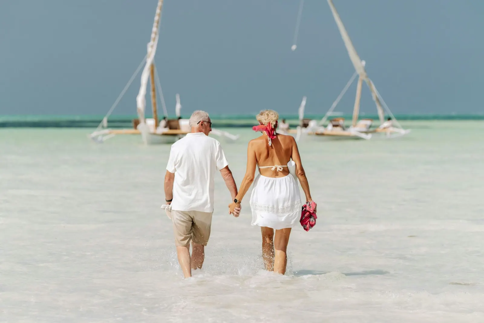 A couple walking hand in hand through turquoise shallows with dhows at anchor behind them